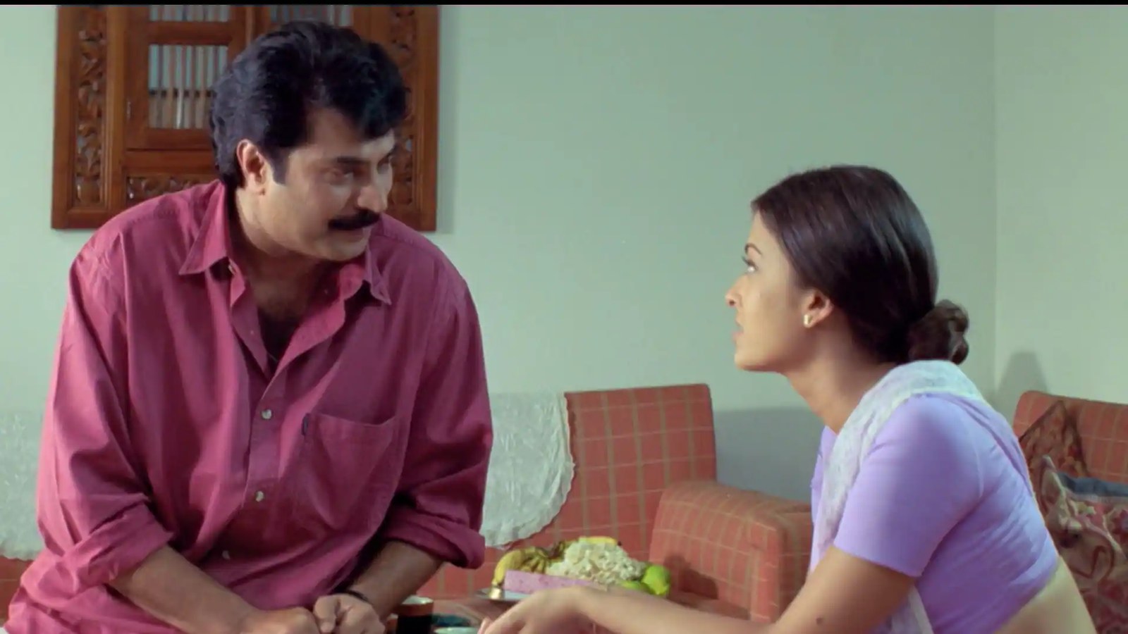 Major Bala sitting across from Meenakshi in a living room, his eyes lowered, hers fixed on him